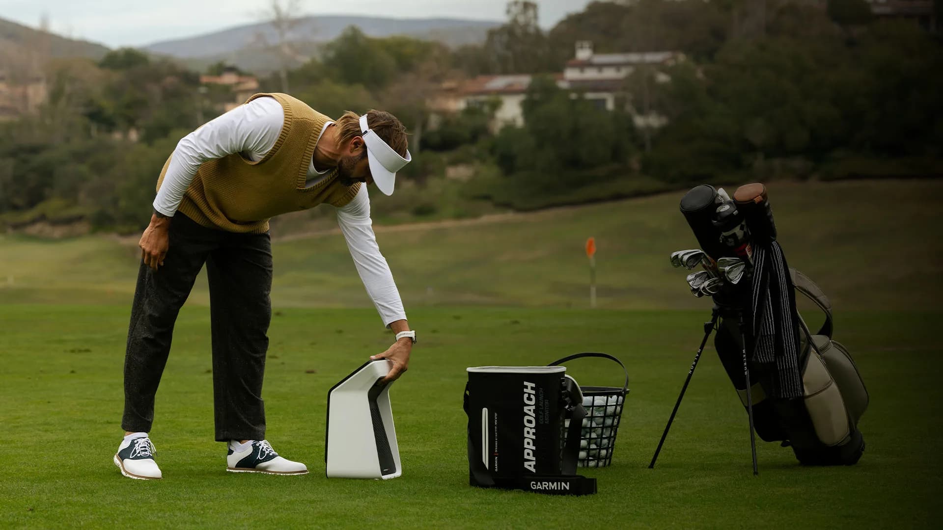 Golfer in a yellow vest positions a white Garmin launch monitor on a green fairway.
